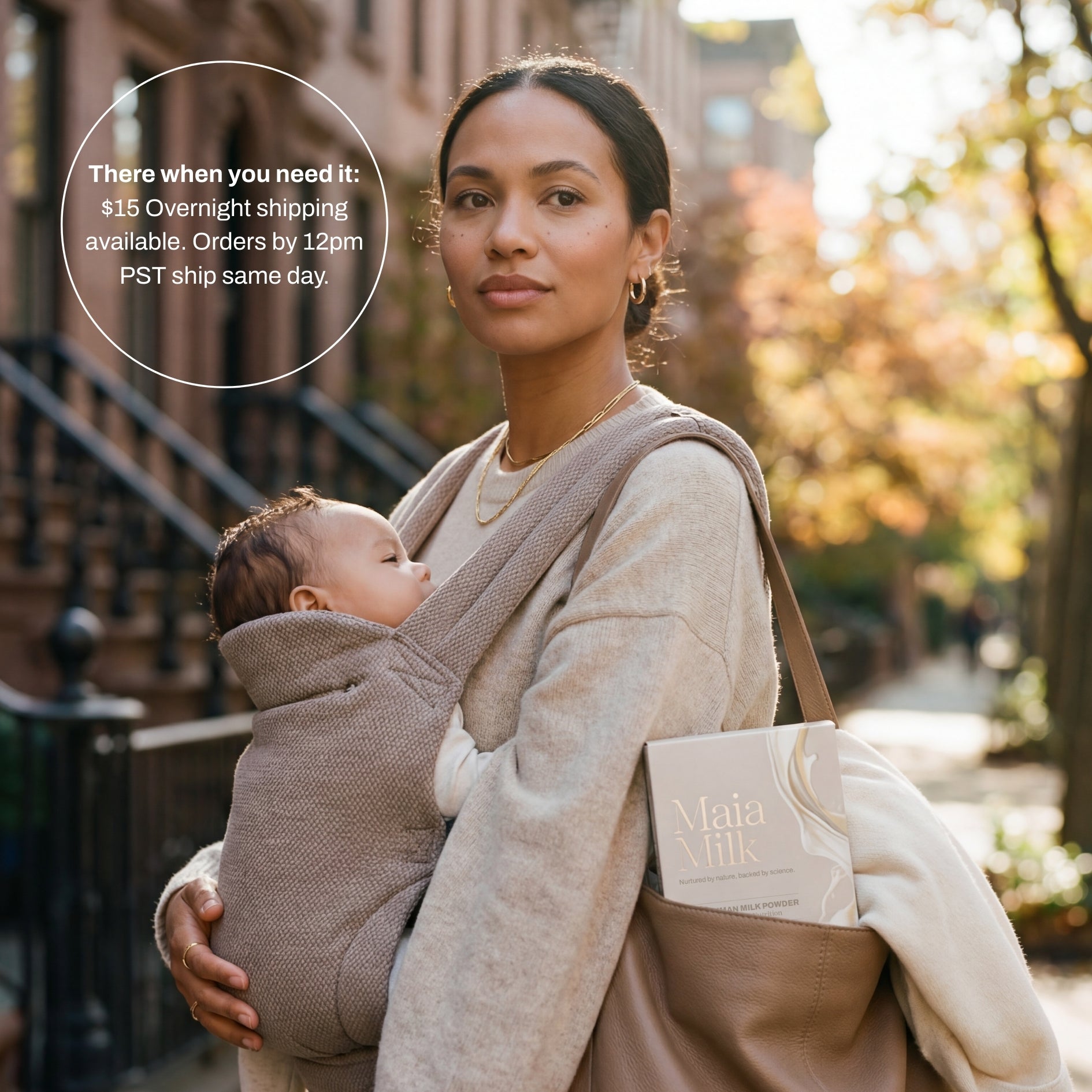 Woman holding a baby in a sling with a MaiaMilk box peeking out of her bag on a city street.